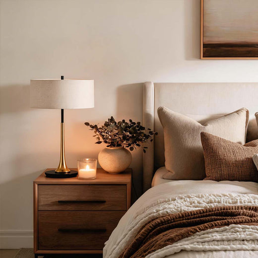 Modern gold fabric table lamp with a cream linen shade on a wooden nightstand in a cozy bedroom.