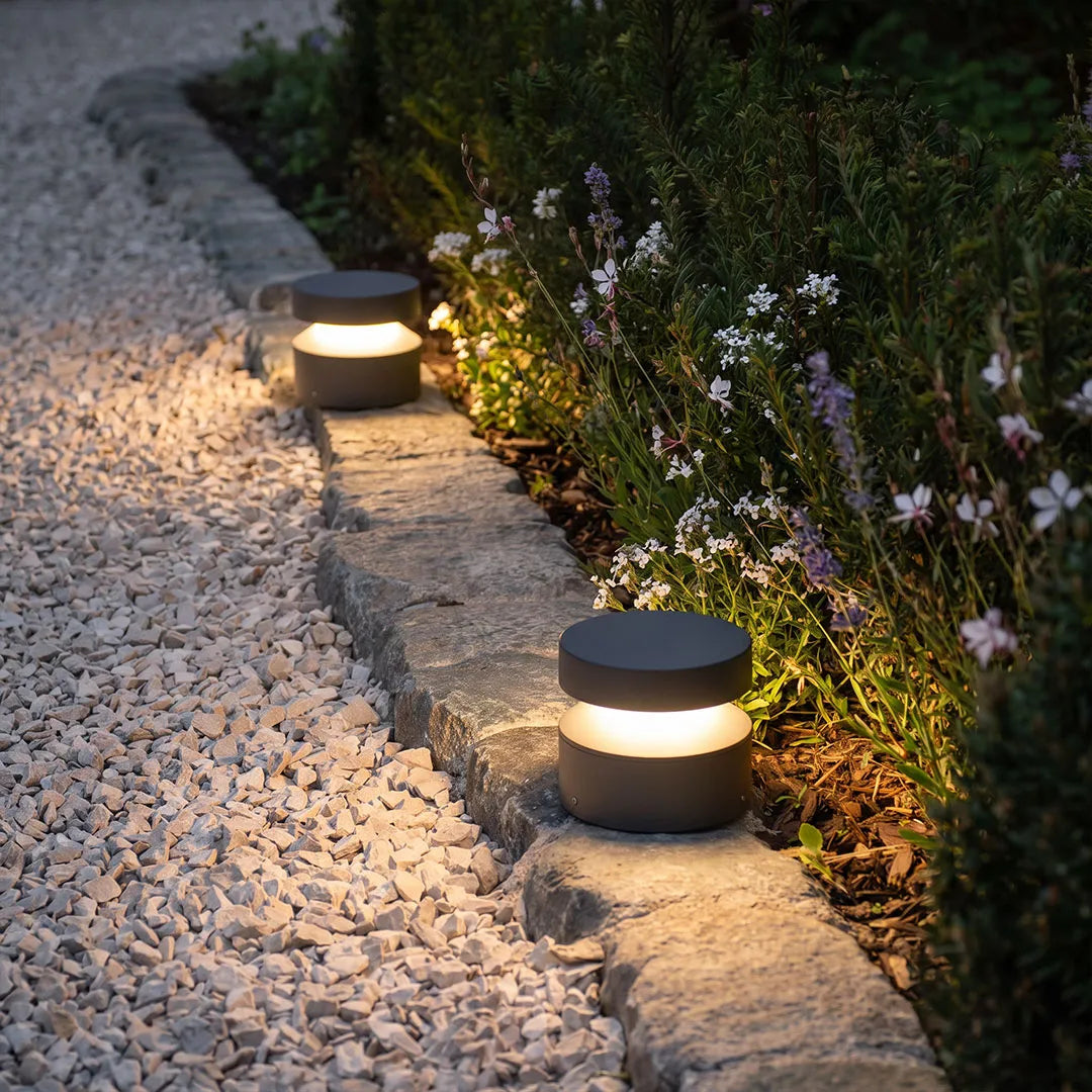 110v landscape lights installed along a stone garden border, providing warm pathway illumination beside flowers and gravel.