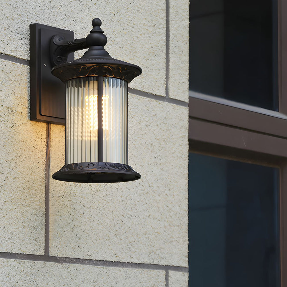 Black outdoor lantern mounted on concrete wall showing warm LED lighting through ribbed glass shade
