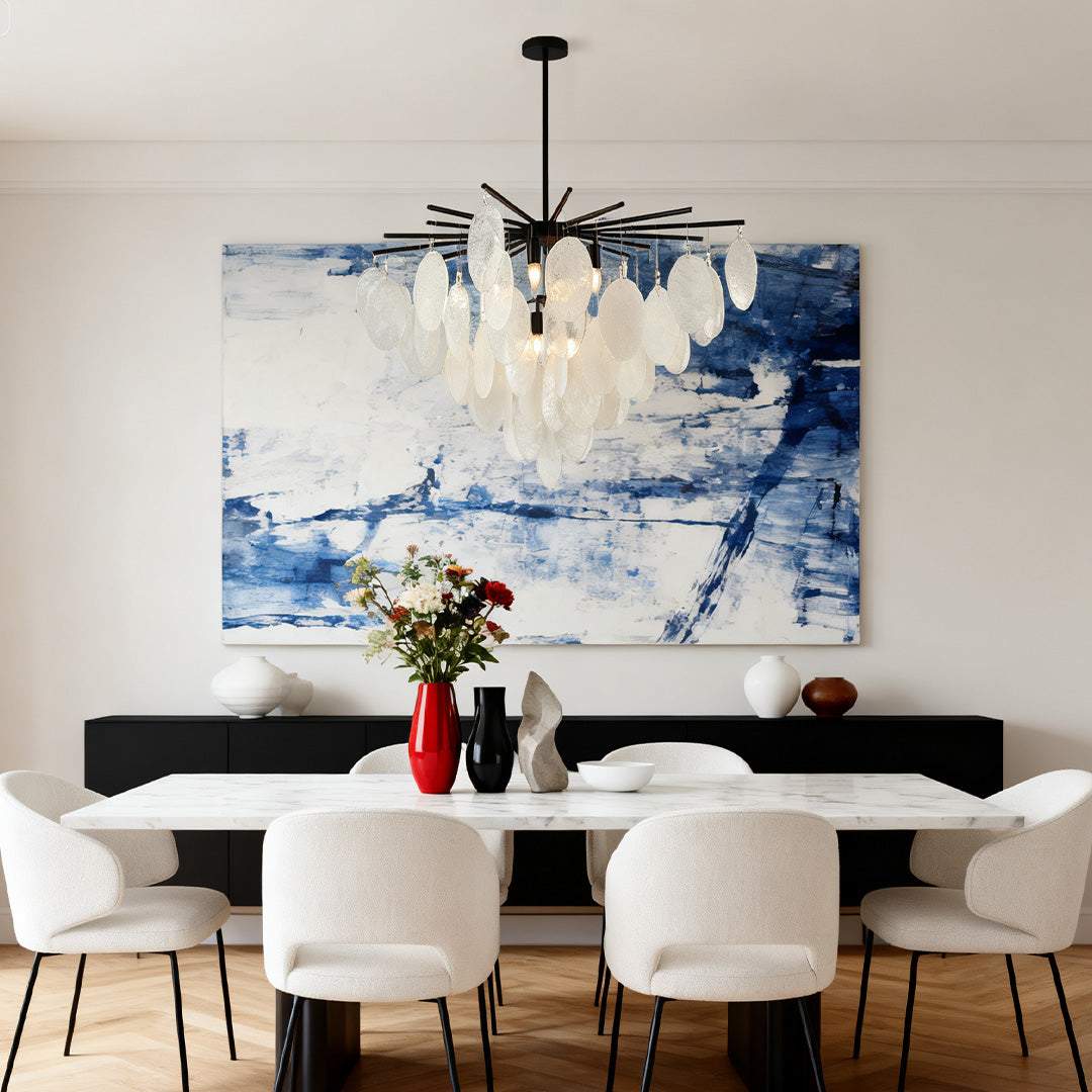 Mother of pearl chandelier suspended over a white dining table, complementing a large abstract blue and white wall art piece.