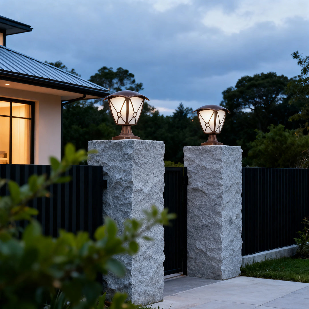 Pair of aluminum pillar light glowing warmly on grey concrete columns flanking black metal gate