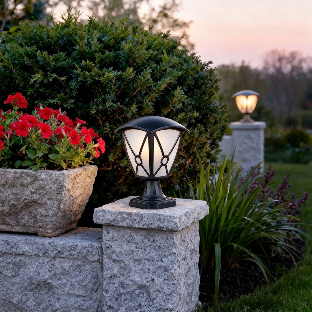 Aluminum pillar light mounted on a square stone column beside flowers and trimmed shrubs.