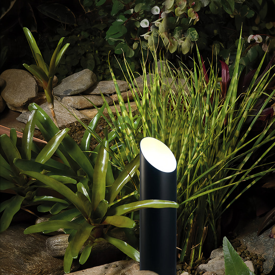 Close-up of the angled top of a metal landscape tree up lighting fixture nestled among vibrant green tropical plants and rocks.