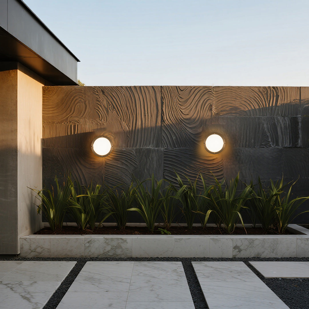 Architectural garden feature lit by two modern flush round outdoor wall lights above low-growing plants.