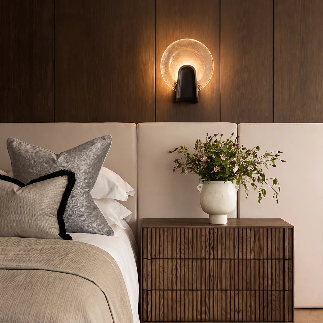 Art deco wall sconce lighting with a round smoky gray glass shade and black finish above a bedroom nightstand.