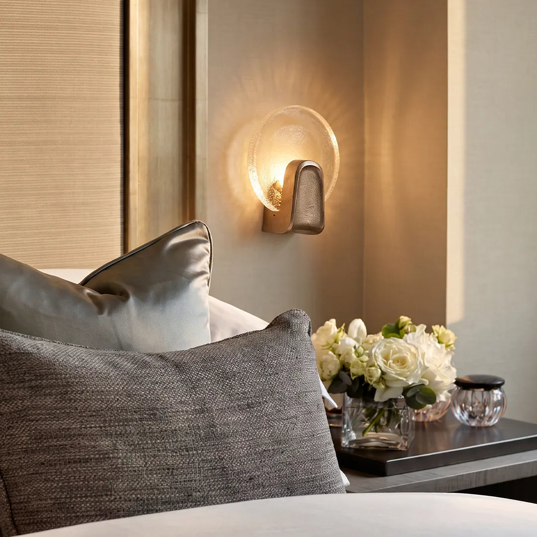 Art deco wall sconce lighting with a round glass shade beside a bed in a warm neutral bedroom.