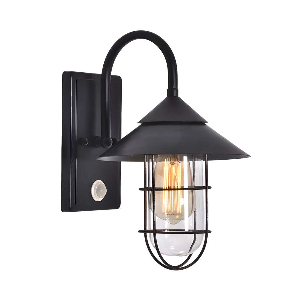 Black barn wall light with curved arm and clear glass, mounted on a brick wall beside a window.