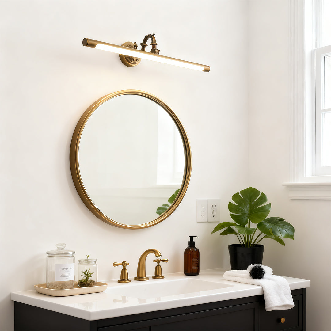 Bathroom LED vanity light mounted above round brass mirror in white bathroom with plant accent.