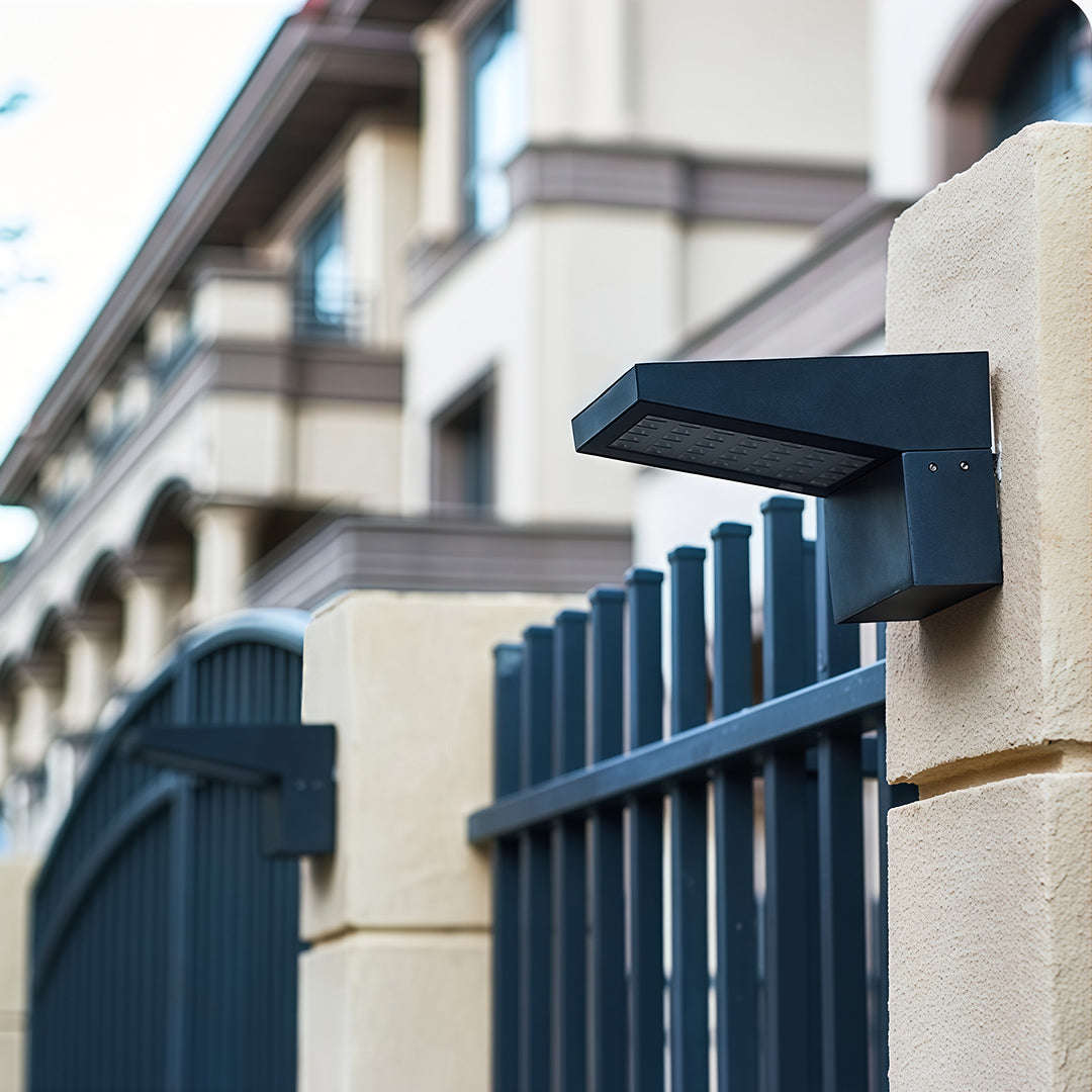 Black angular wall mount outdoor light fixtures installed on a gate pillar next to a metal fence.
