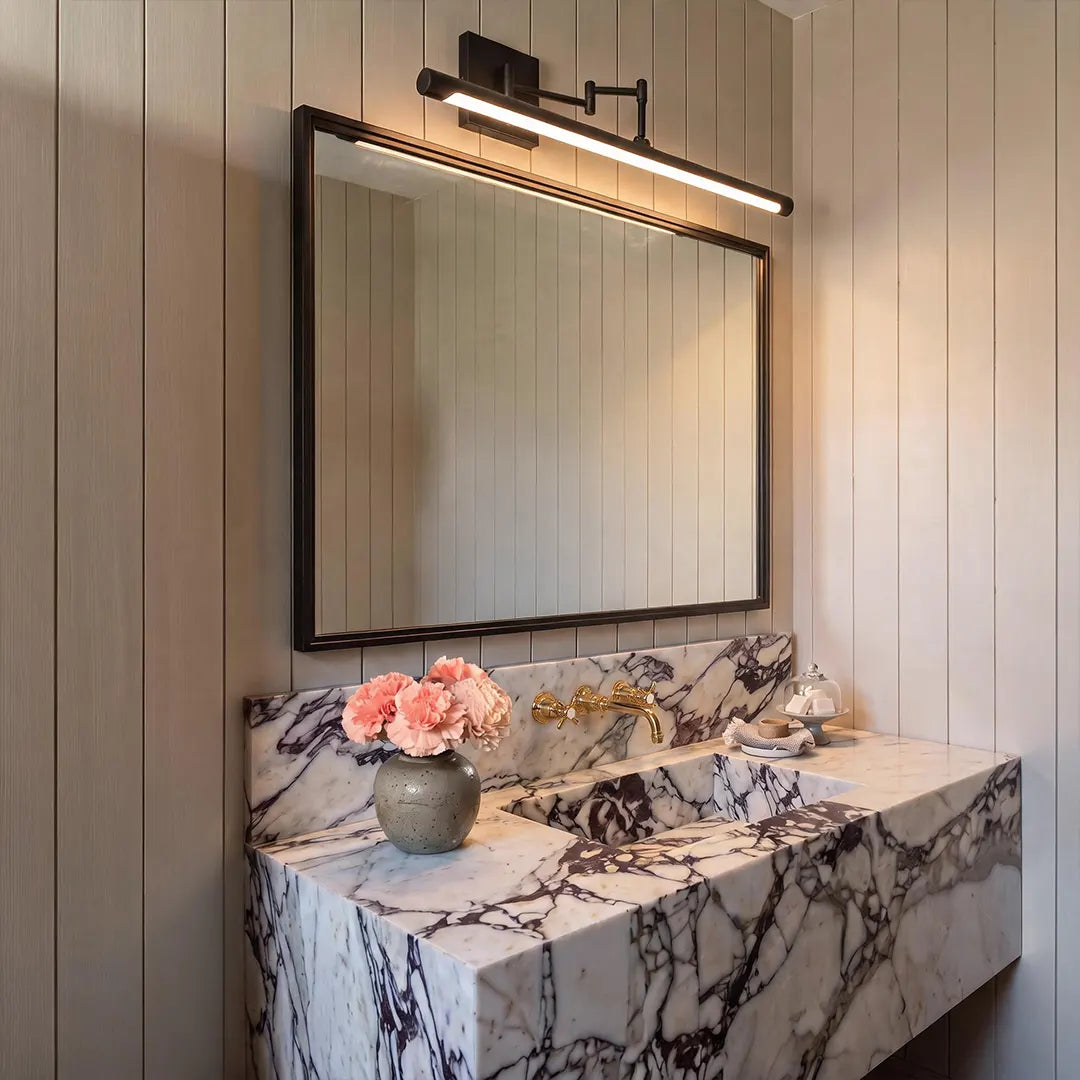 Black bar vanity light mounted above a rectangular bathroom mirror with a marble vanity and warm neutral wall paneling.