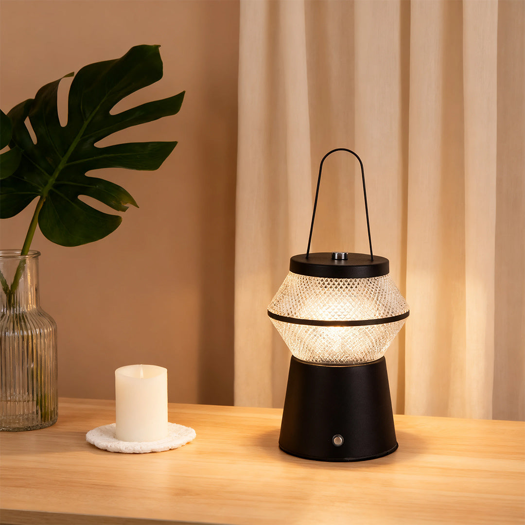 Black base led outdoor solar lantern illuminating a modern indoor desktop next to a large potted monstera leaf. 