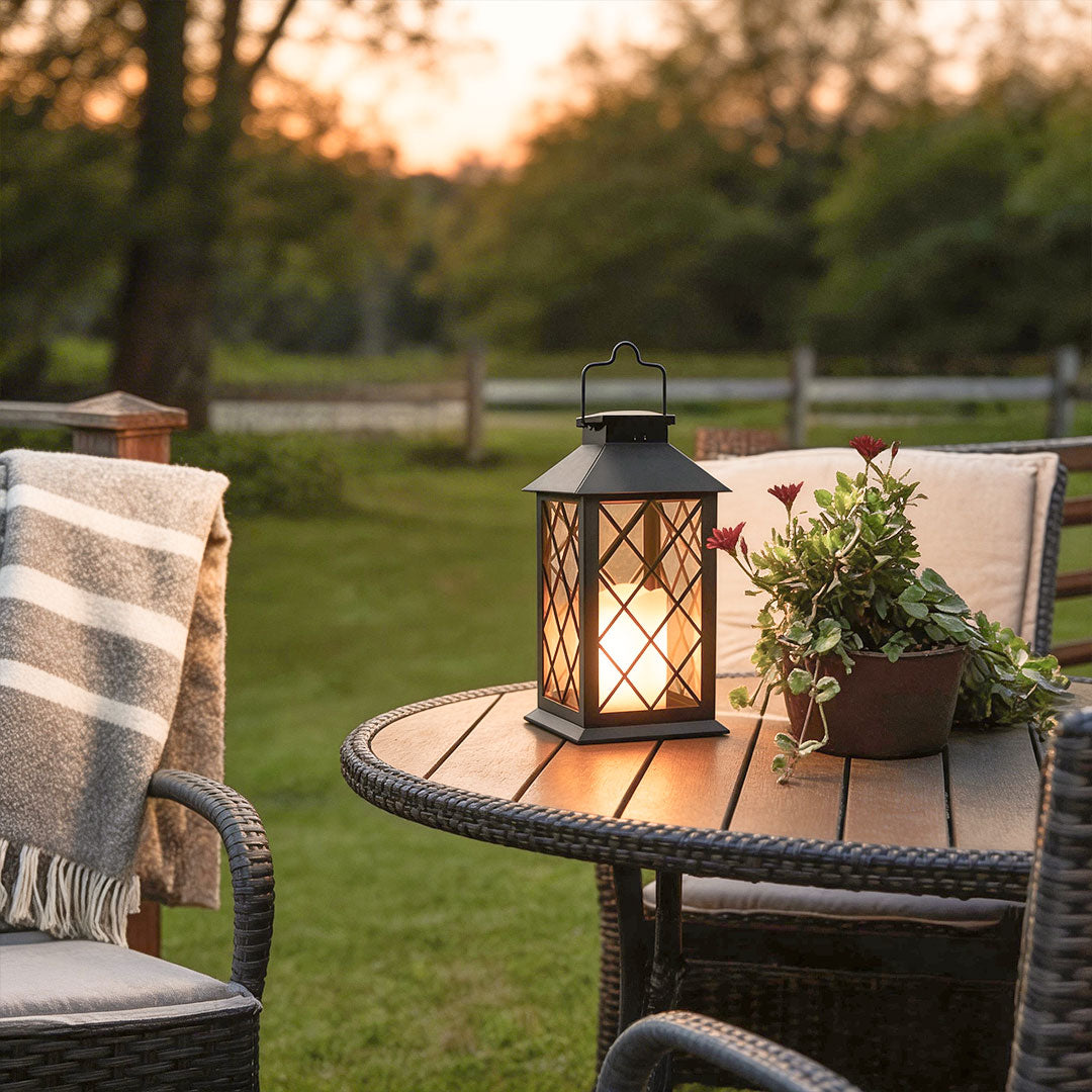 A black candlestick lantern glowing on a patio table at sunset with a cozy blanket and flowers.