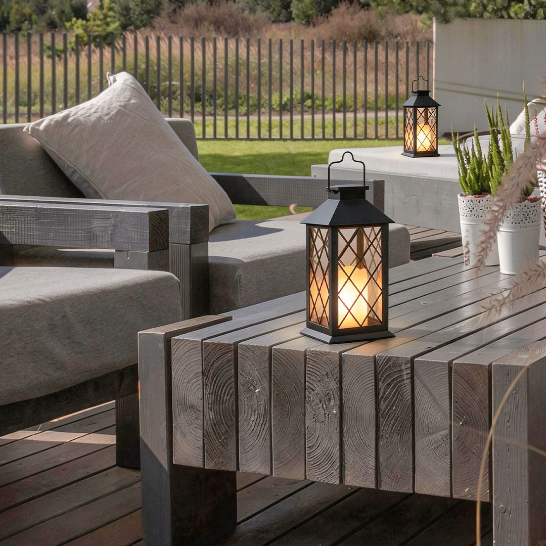 Black candlestick lantern glowing on a rustic wood patio table with garden view in the evening.