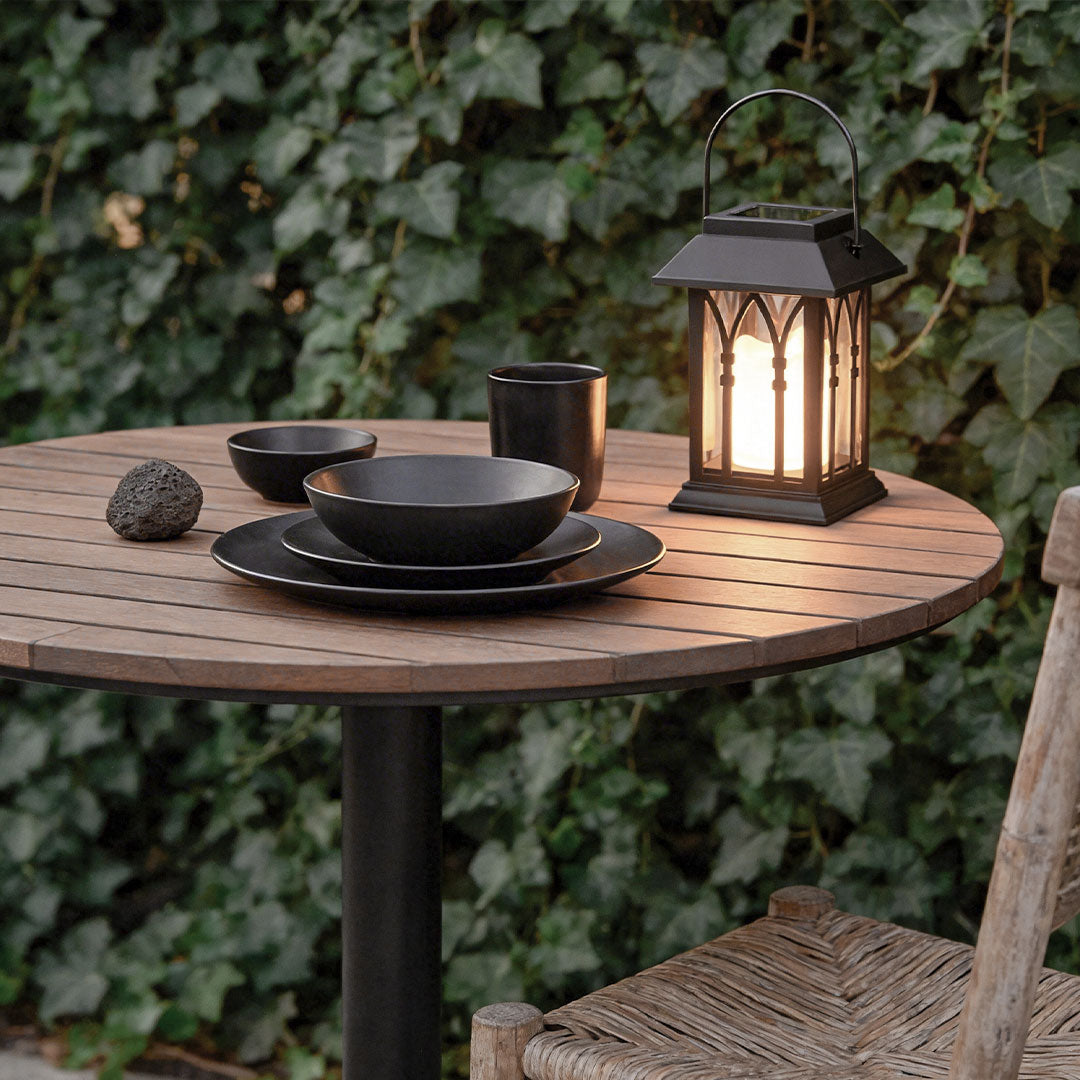 A black candlestick lantern on a wooden dinner table with black plates and ivy leaves behind.