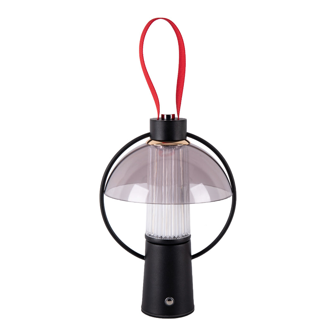 Isolated view of a black circle table lamp with a clear shade and a contrasting red fabric handle for portability. 