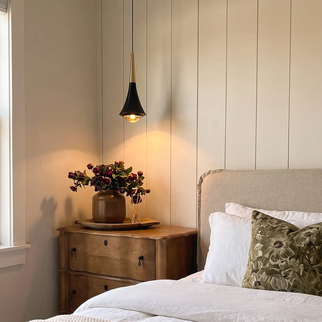 Black cone pendant light suspended beside a bed, casting warm light in a cozy bedroom setting.