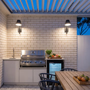 Black cone wall sconce pair illuminating an outdoor kitchen and dining area beneath a pergola.