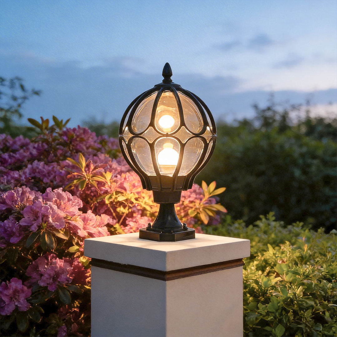 Black outdoor globe pillar light glowing on a white stone post with purple flowers at twilight.