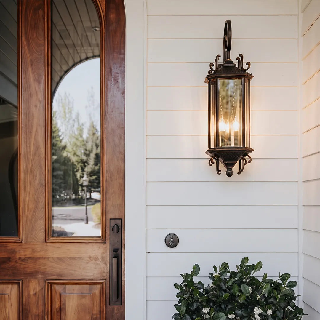 Black outdoor lantern sconce fixed beside a wooden front door, highlighting the entrance with warm candle-style lighting.