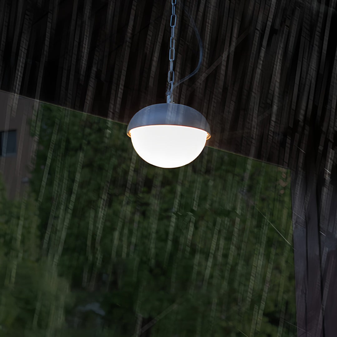 Black outdoor pendant light glowing softly at night, suspended against a backdrop of greenery.