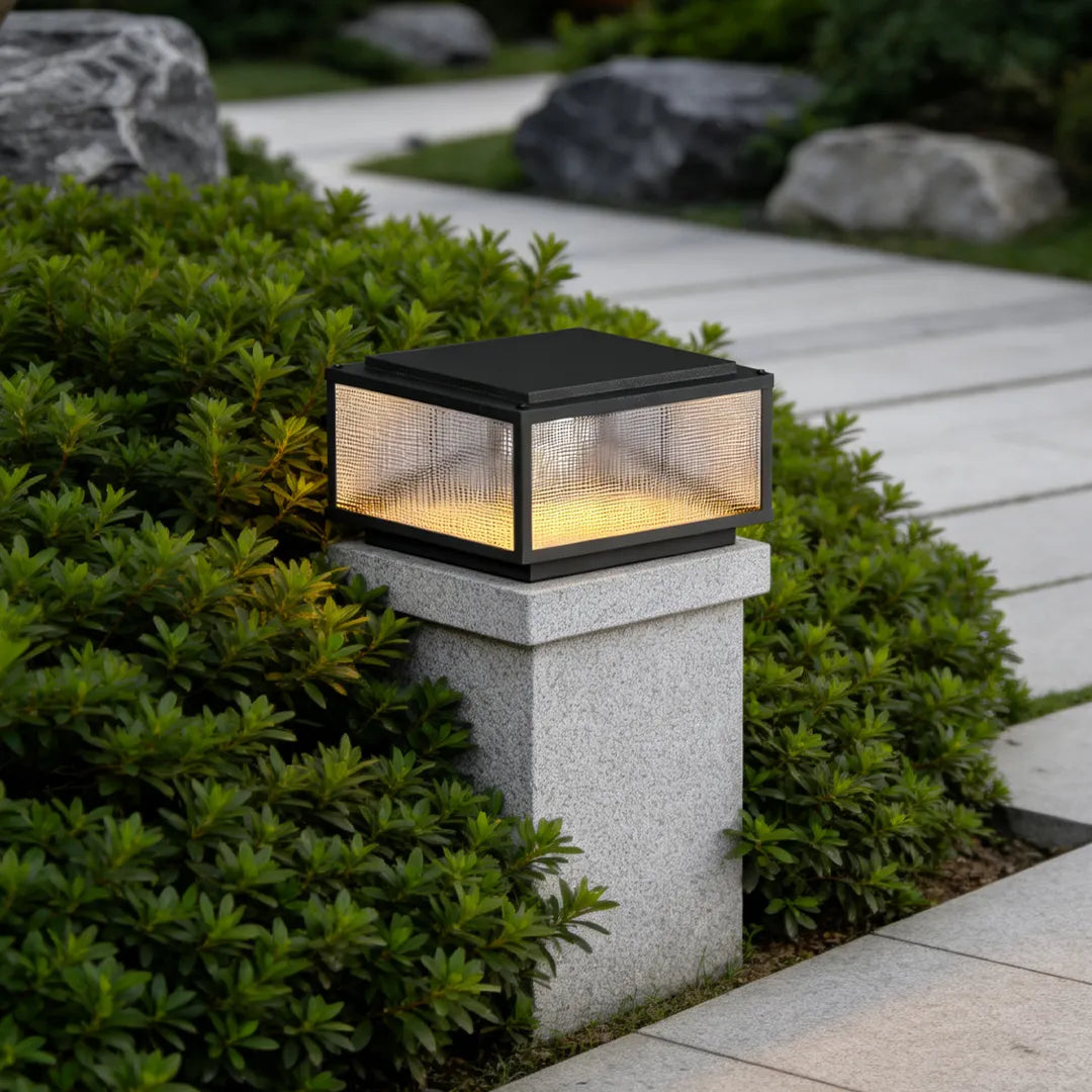 Black outdoor pillar light fixture installed on a square post beside shrubs and a garden path.