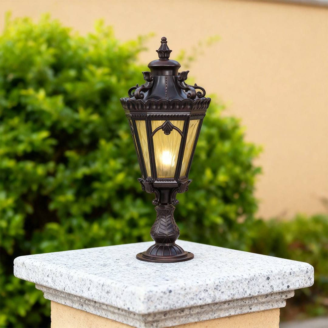 Black outdoor pillar light glowing warmly on granite post beside green plant against beige wall