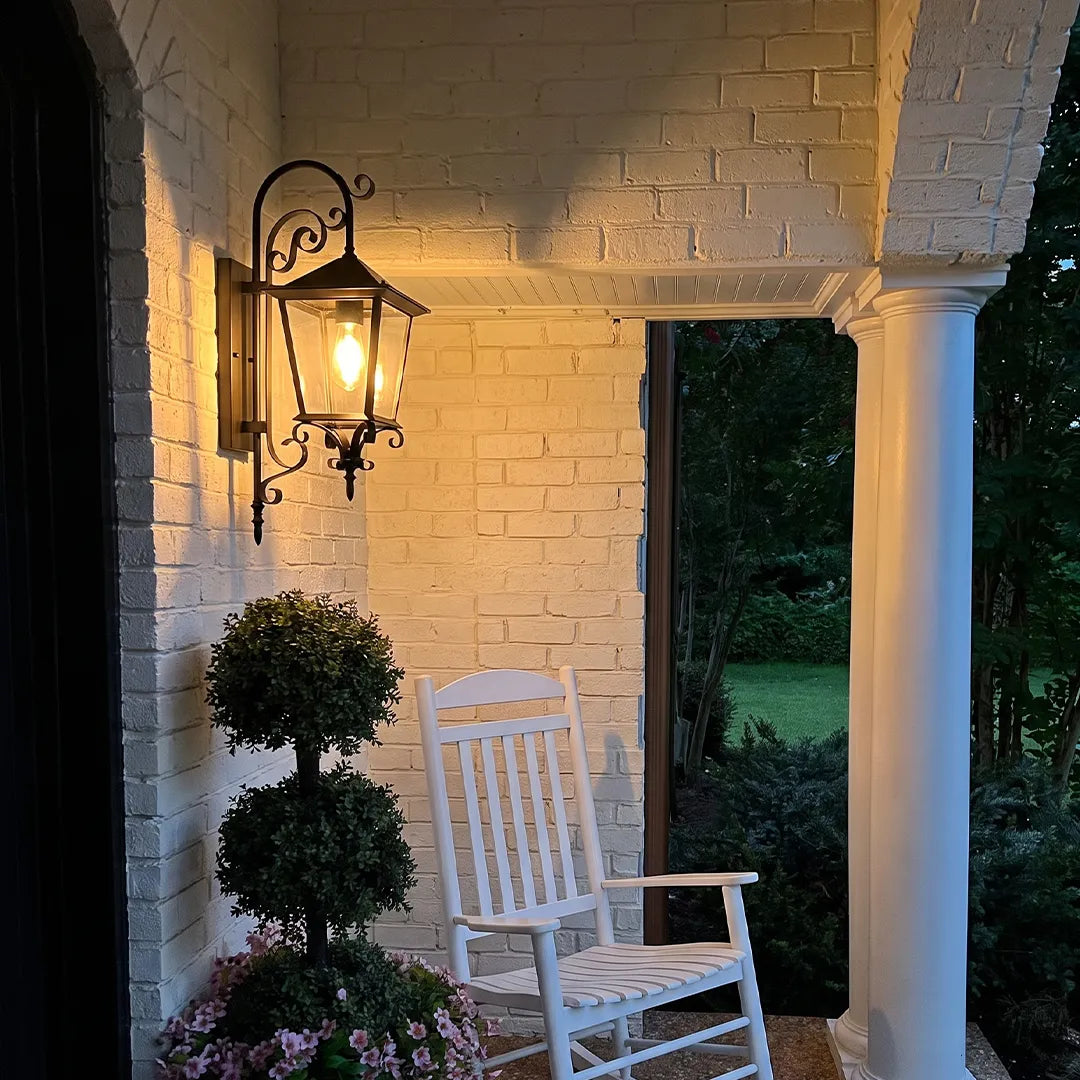 Black outdoor sconce beside a porch column at dusk, casting a warm glow across the white brick exterior.