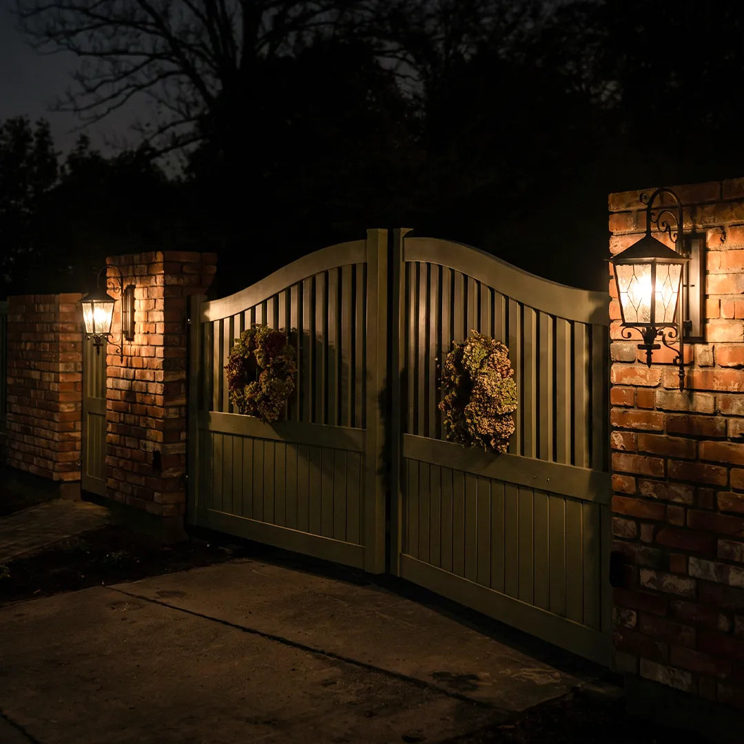 Black outdoor sconces mounted on brick gate columns at night, creating an elegant and welcoming driveway entrance.