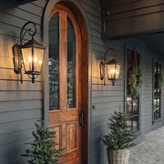 Black outdoor wall light mounted beside a tall arched wood front door, bringing classic lantern-style illumination to the entryway.