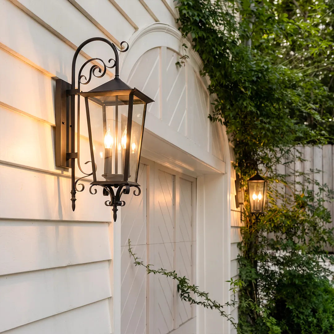 Black outdoor wall sconce mounted on white siding beside an arched garage door, featuring a traditional lantern design with scroll arm details.