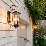 Black outdoor wall sconce mounted on white siding beside an arched garage door, featuring a traditional lantern design with scroll arm details.