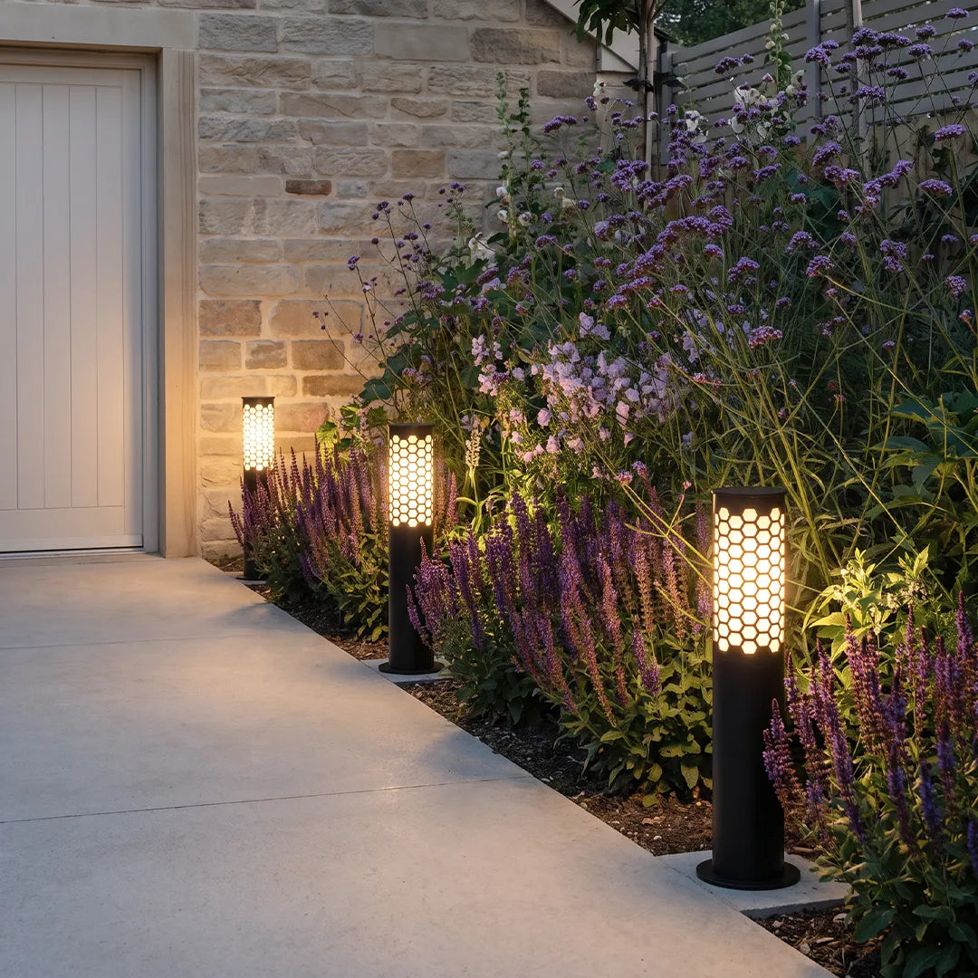 Black perforated outdoor bollard lights illuminating a flower bed beside a modern driveway