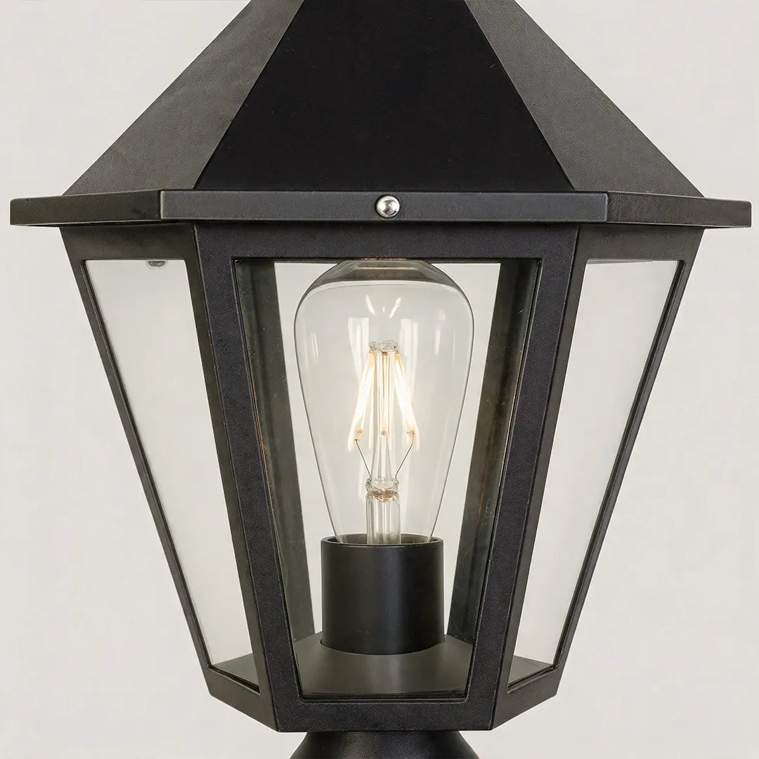 Black pillar light close-up showing the tapered lantern frame, clear glass panels, and exposed bulb detail.