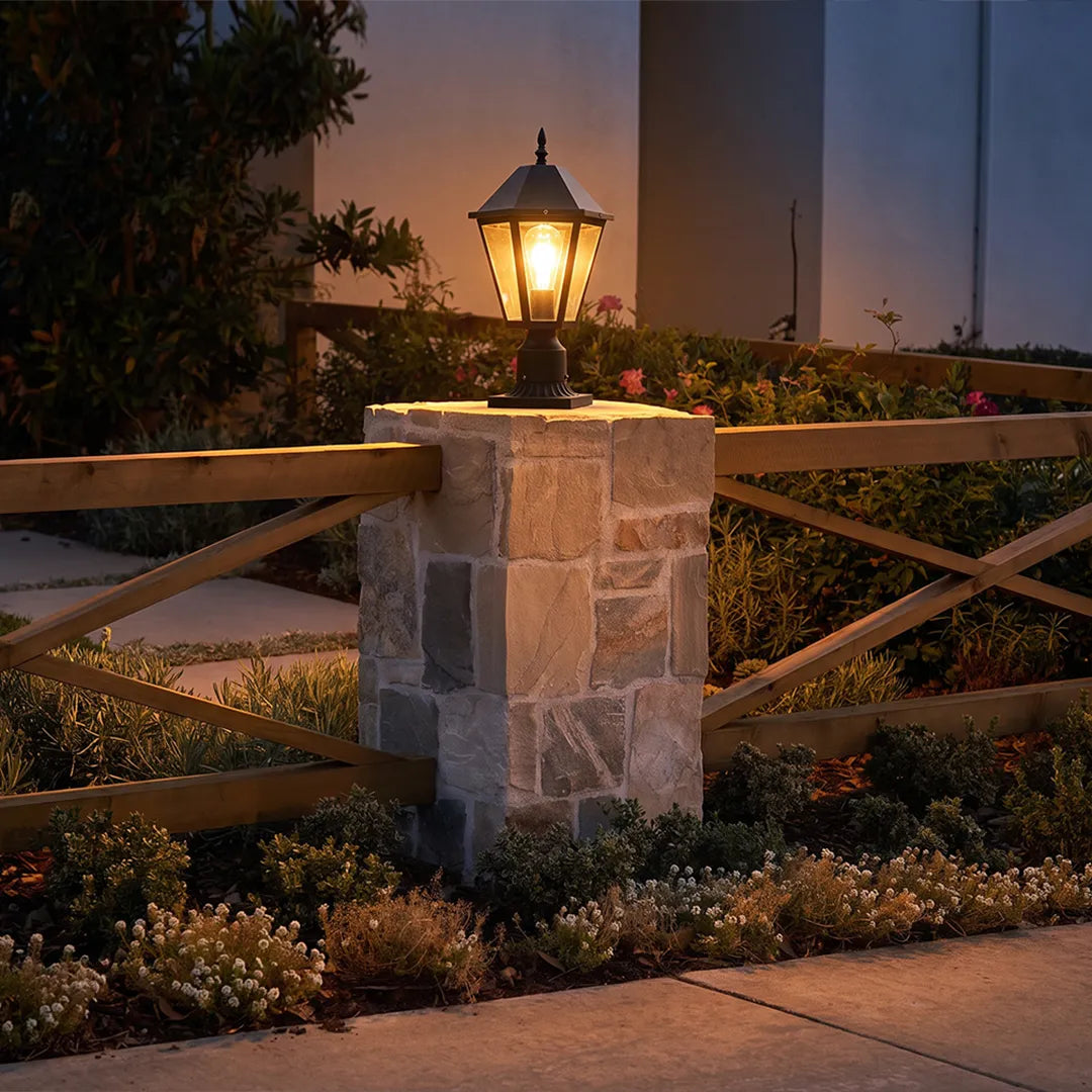Black pillar light placed on a stone entry post at dusk, illuminating the driveway with a warm lantern glow.