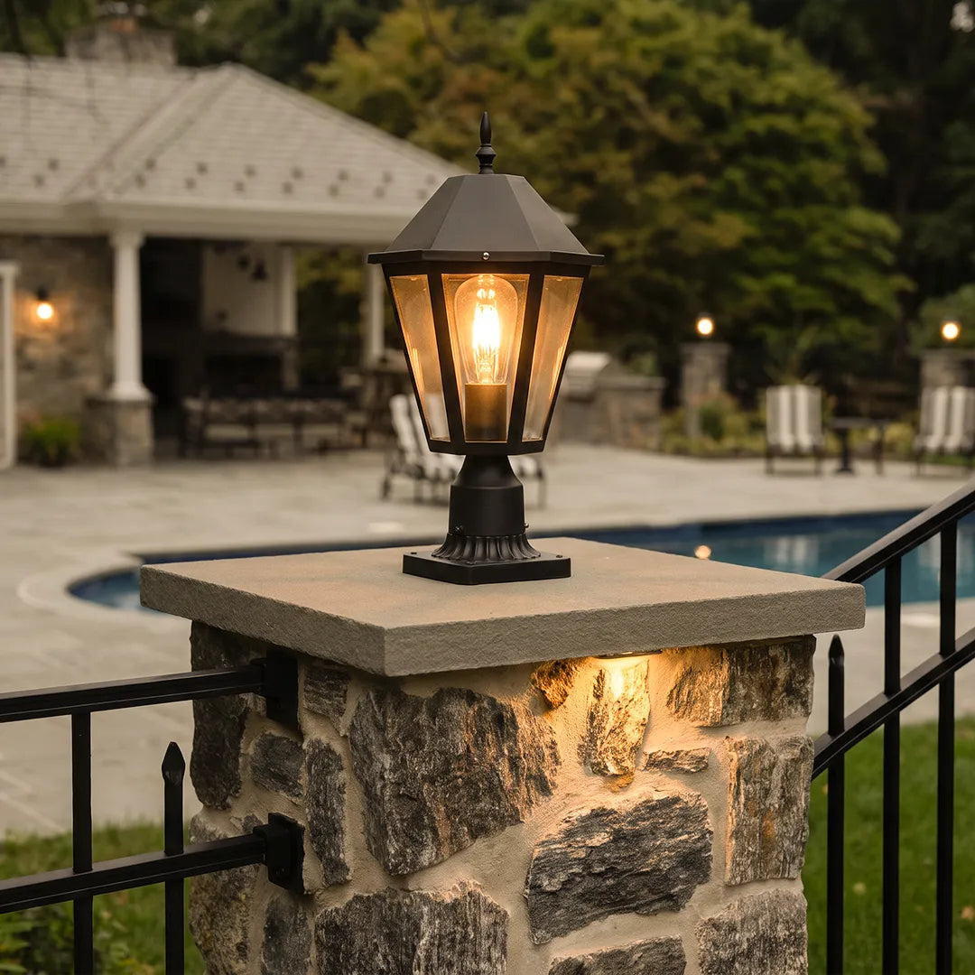 Black pillar light mounted on a stone gate post beside a poolside patio, adding warm traditional outdoor illumination.