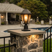 Black pillar light mounted on a stone gate post beside a poolside patio, adding warm traditional outdoor illumination.