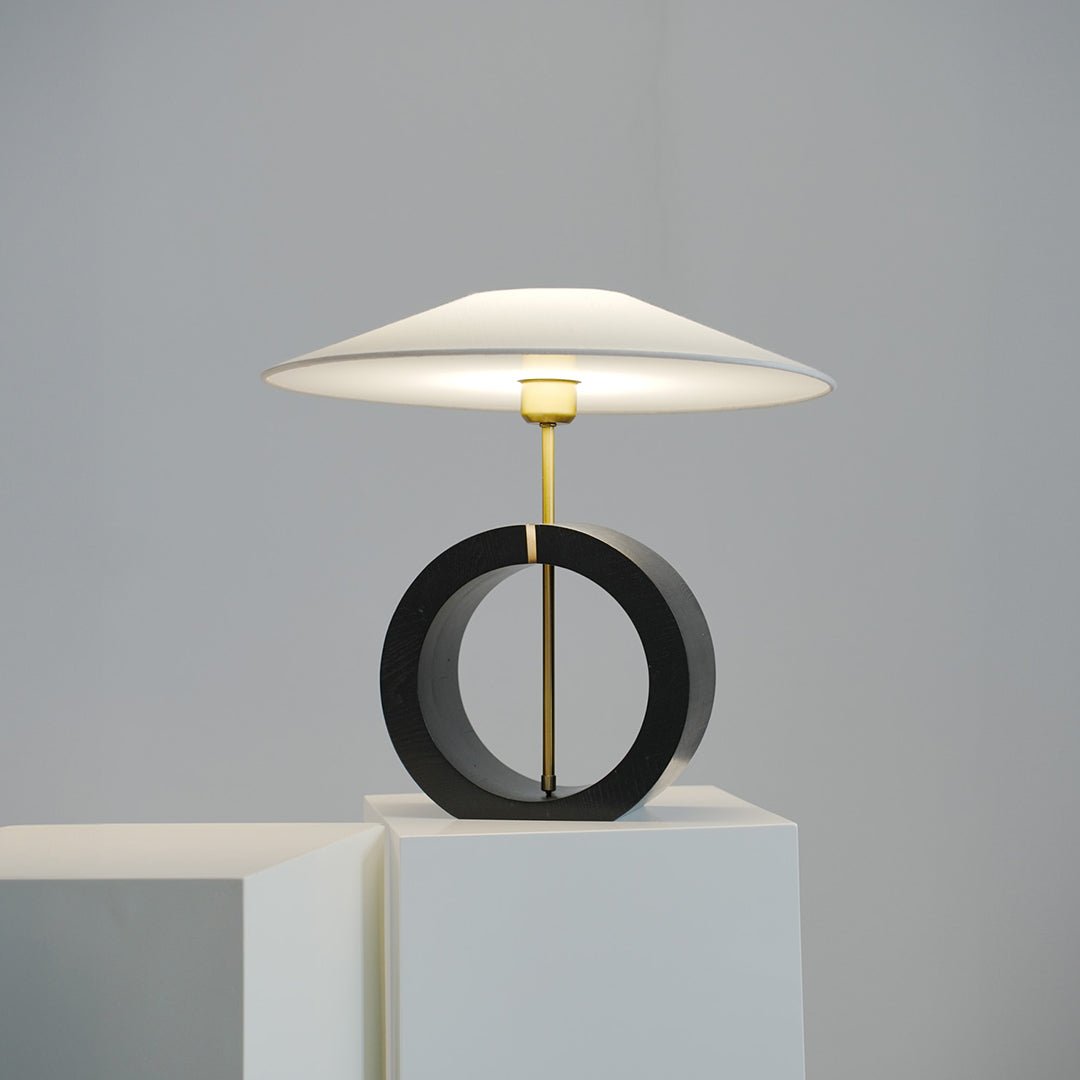 Black circular base table lamp with white shade placed on modern display pedestal.