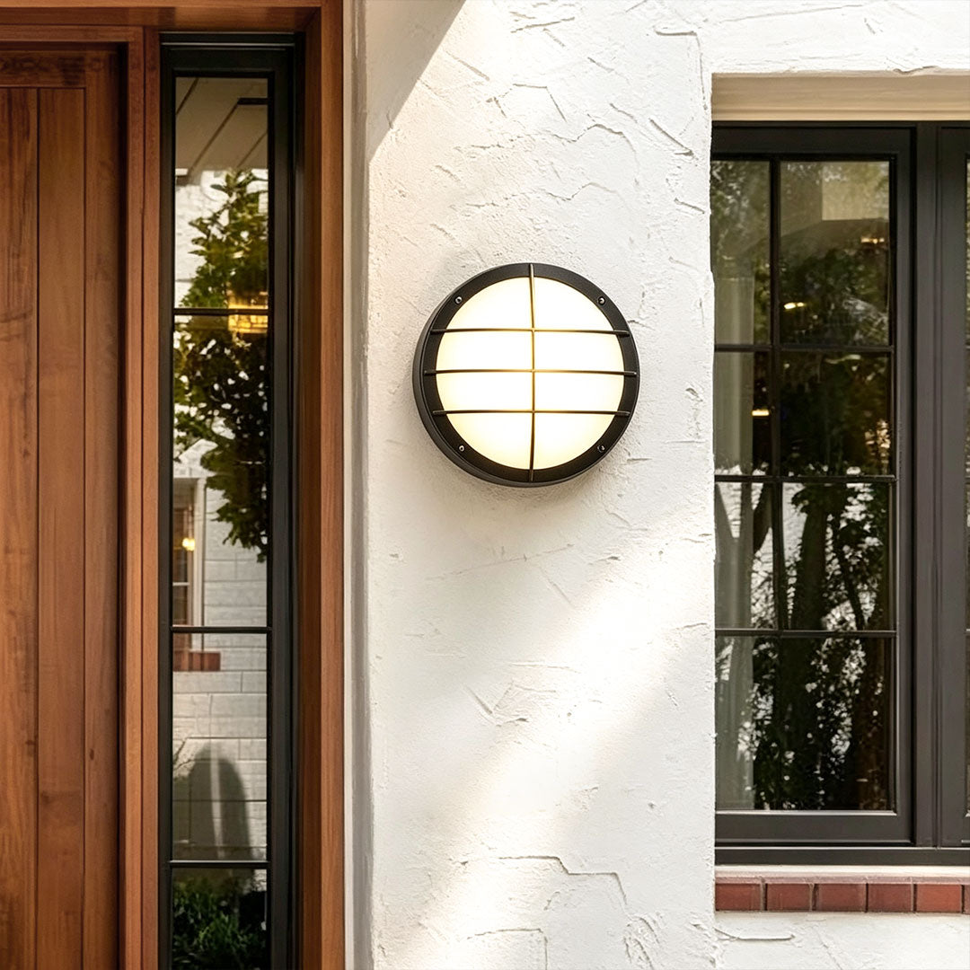 Black round outdoor wall light fixture with grille detail mounted on a white exterior wall near a front door.