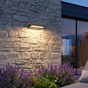 Black sconce wall lights illuminating a textured stone exterior wall beside purple garden flowers.