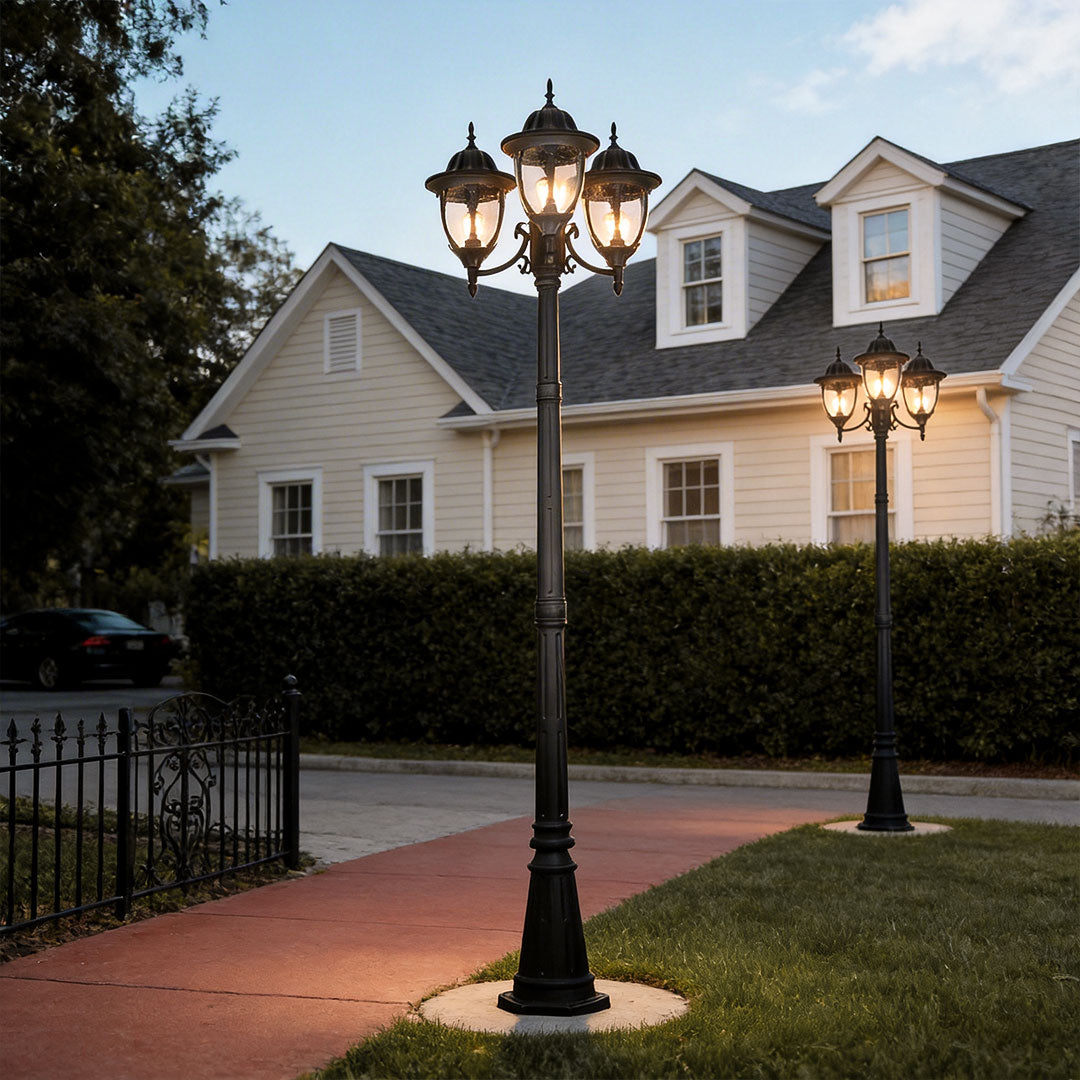 Black three-head street lamp post light glowing at dusk on a sidewalk in front of a cozy house.