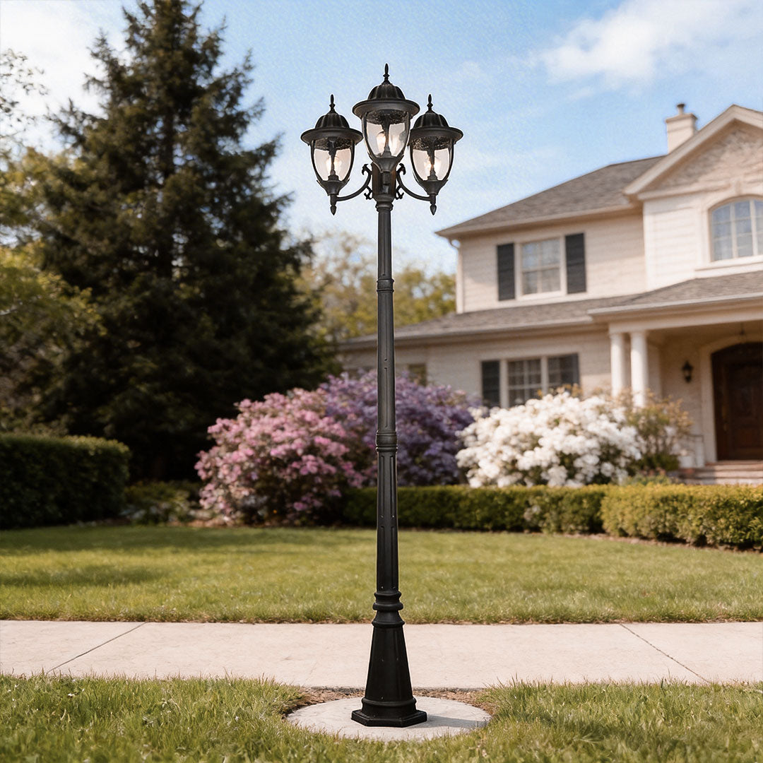 The black three-head street lamp post light in a front yard garden with pink flower bushes.