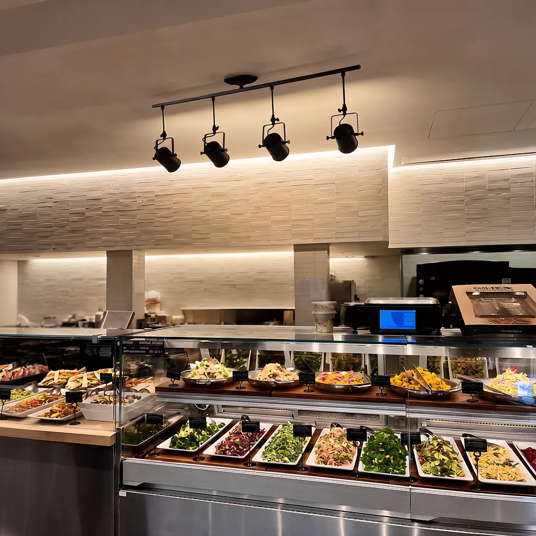 Black track light with adjustable spotlights illuminating a food counter and display area.