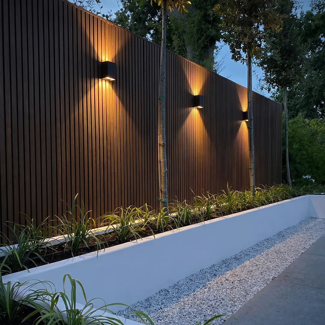 Black wall lights installed along a slatted garden fence, creating warm accent lighting at dusk.