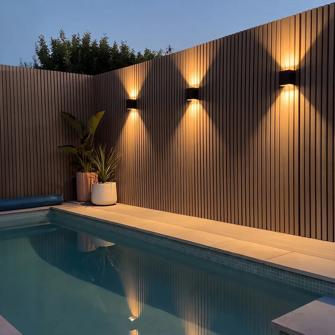 Black wall lights mounted on a curved slatted fence, illuminating a poolside patio with warm beams.