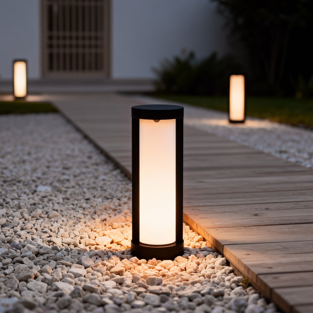 Bollard landscape light with a cylindrical shape, casting warm light on a gravel pathway.