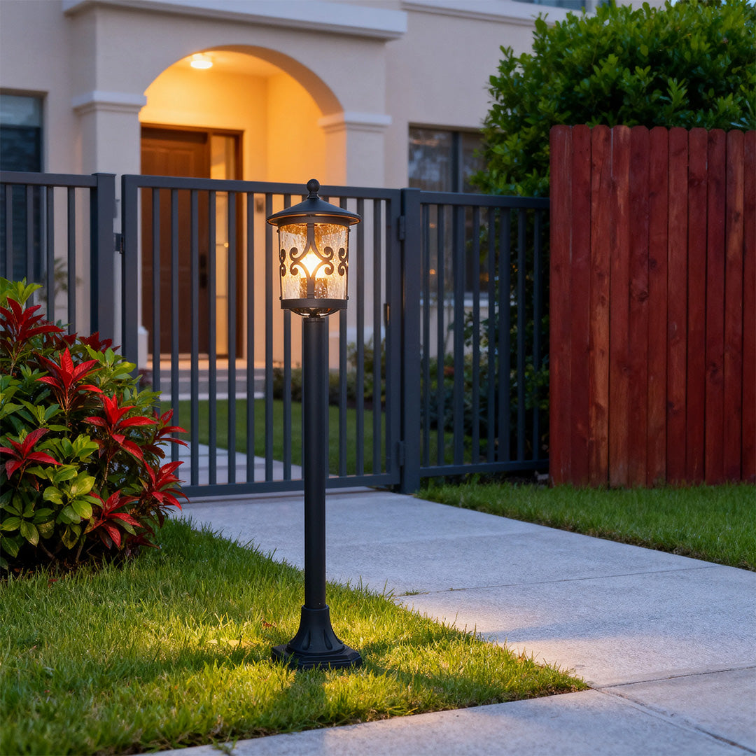Bollard LED light glowing warmly on residential driveway with black metal gate and red flowers