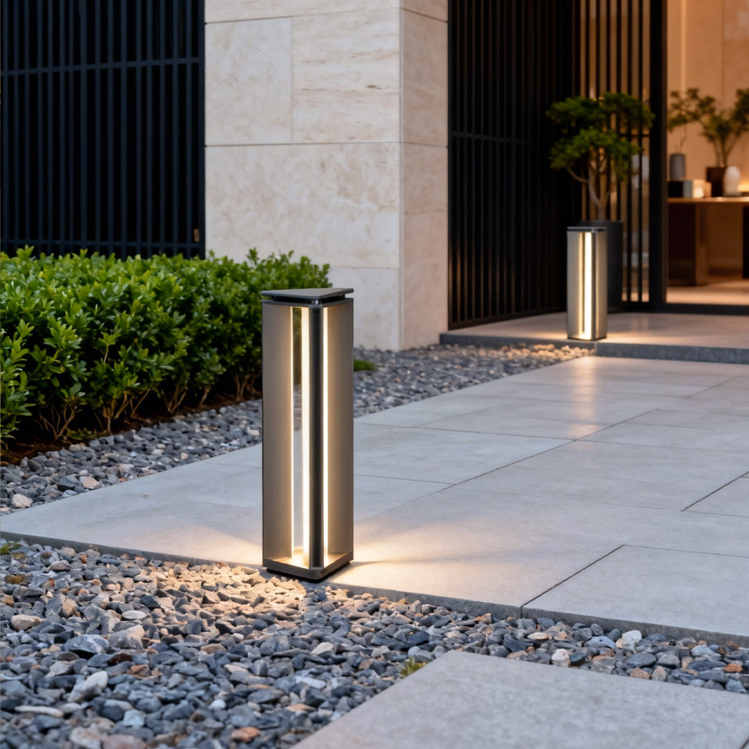 Bronze bollard outdoor lights installed along a walkway beside a building, illuminating surrounding pebbles.