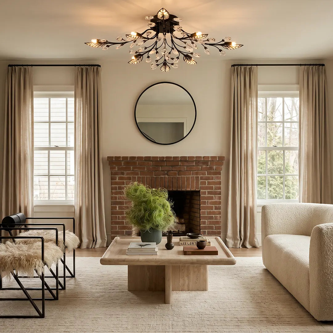 Modern branch chandelier above a living room with a brick fireplace, round mirror, and neutral seating.