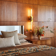 Brass wall sconce light mounted beside a bed on a warm wood accent wall.