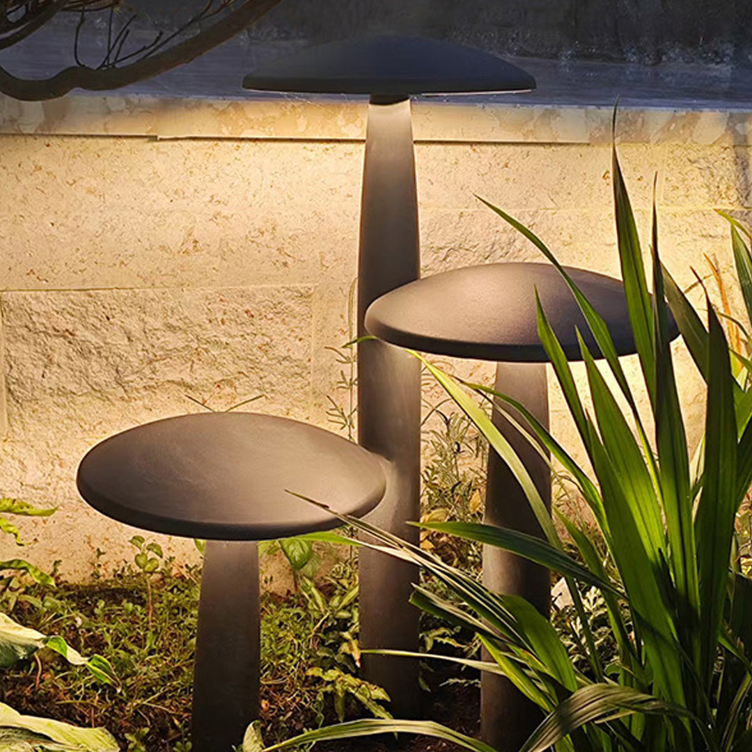Outdoor mushroom bollard light in bronze, enhancing a garden with natural plants and stone walls.